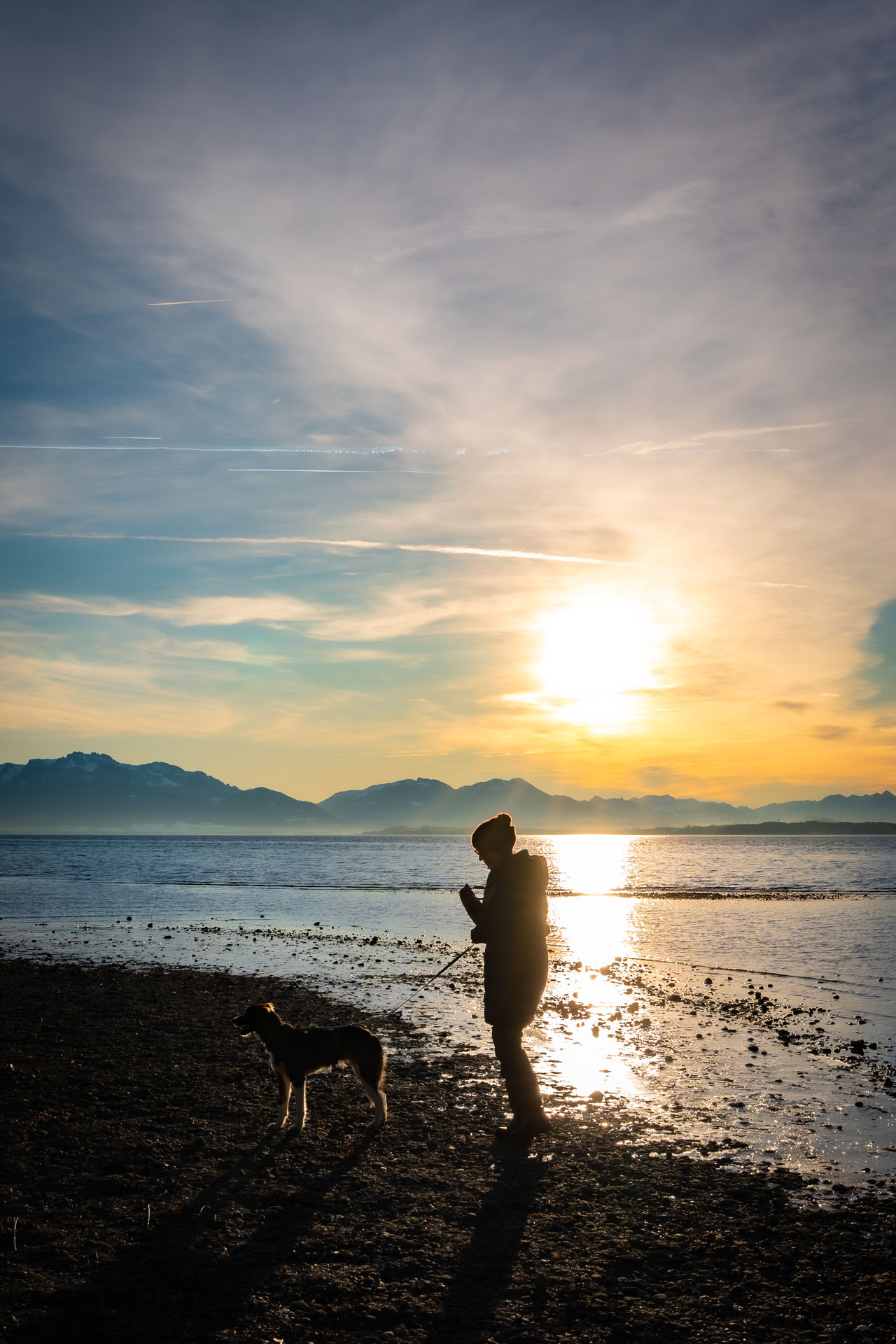 Mensch mit Hund am See – stimmungsvolle Hundefotografie im Gegenlicht Silhouette einer Person mit Hund am Ufer eines Sees bei tief stehender Sonne und Bergpanorama im Hintergrund